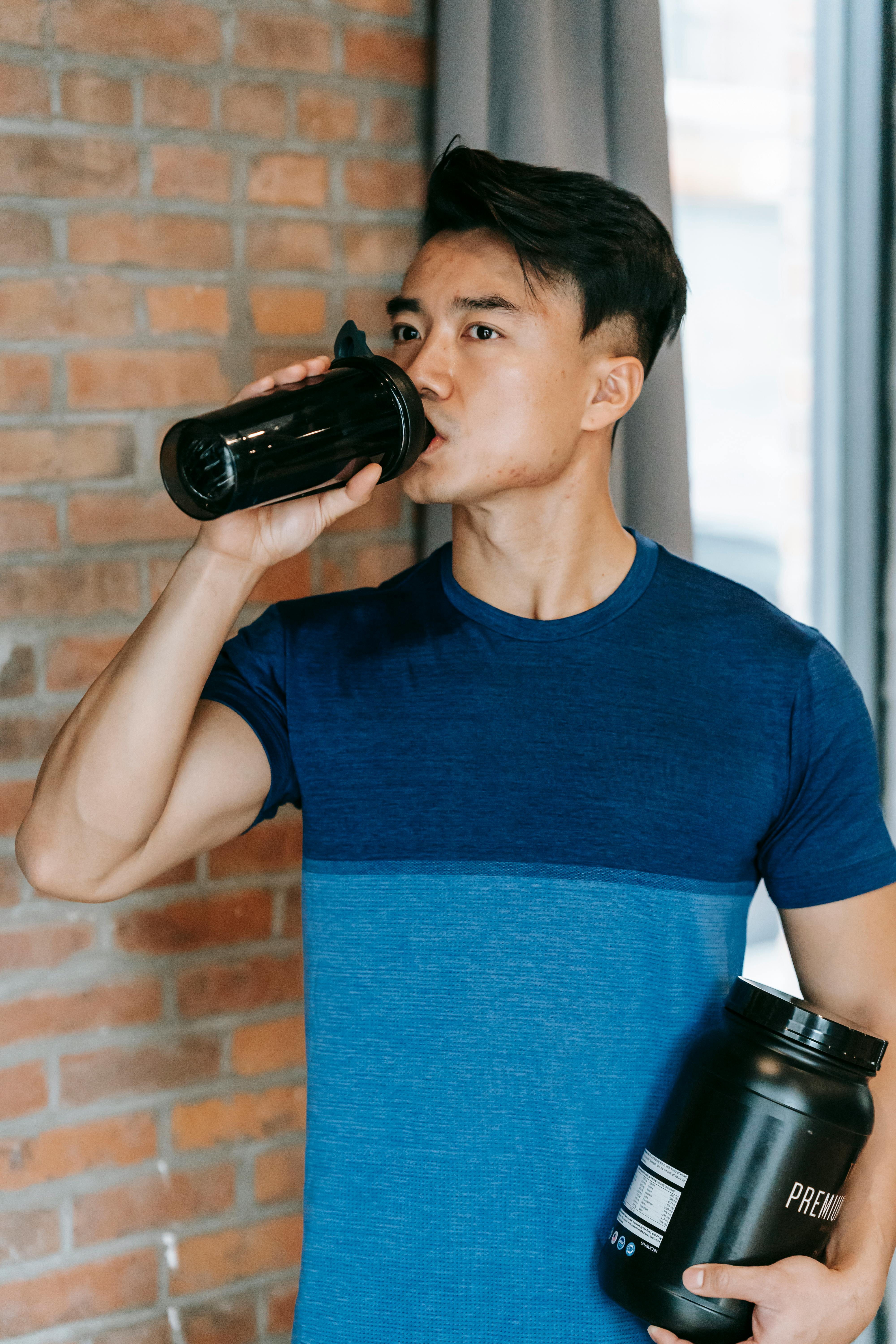 determined young ethnic guy drinking water during training in gym