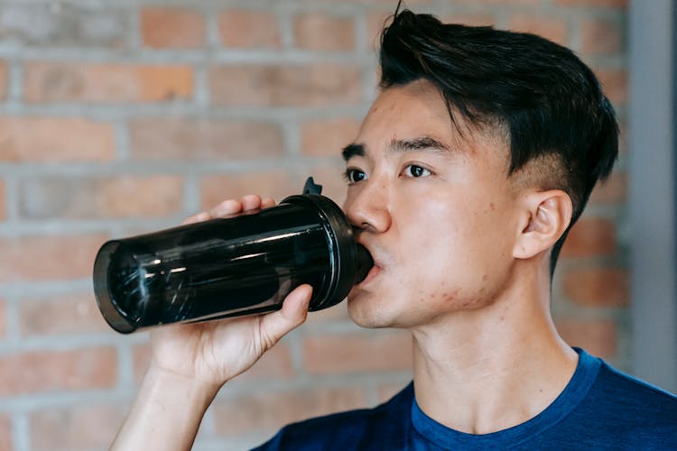 Focused Young Asian Sportsman Drinking Water From Shaker In Gym