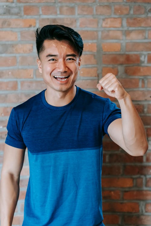Free Cheerful young ethnic male athlete in sportswear smiling and showing fist while standing against brick wall after successful workout in gym Stock Photo