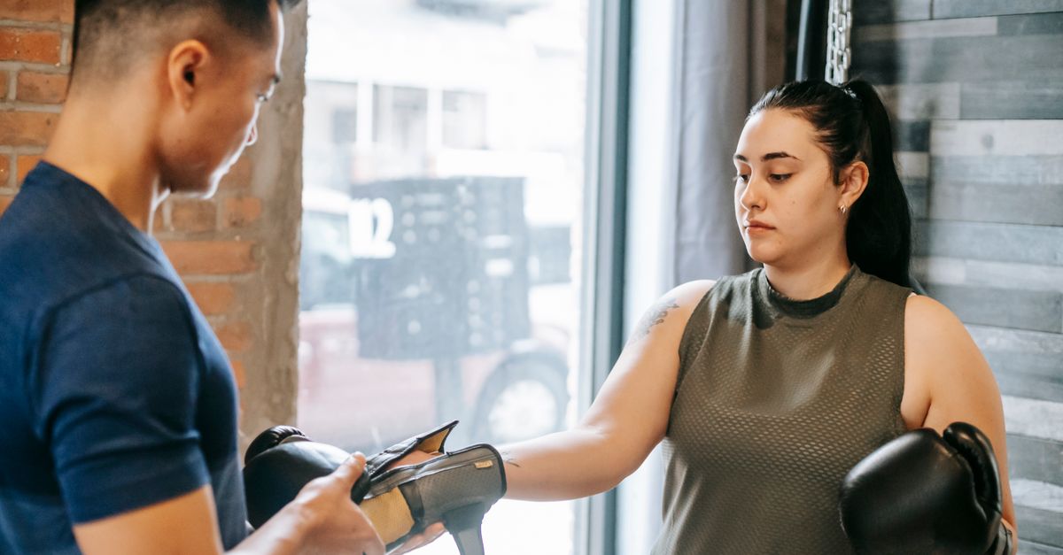 Professional personal trainer preparing focused woman for boxing training while putting on gloves in gym