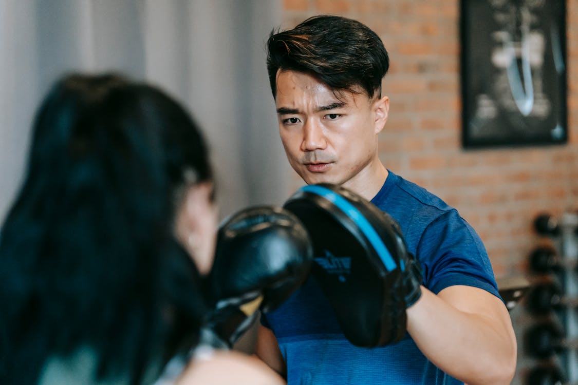 Free Woman in boxing gloves punching paw of concentrated Asian personal instructor during workout in gym Stock Photo