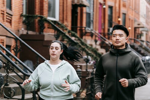 Fit young Asian male trainer with motivated plus size female in activewear running together on city street during weigh loss workout