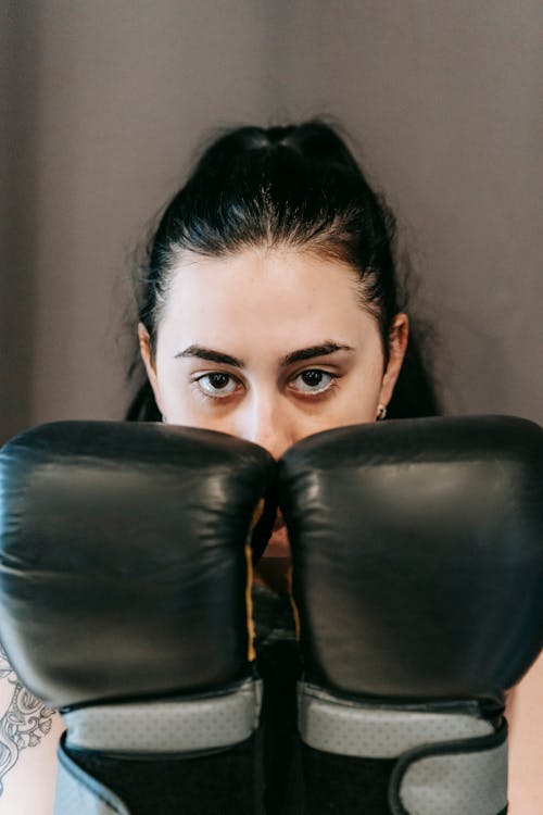 Powerful young sportswoman with long dark hair demonstrating fists in boxing gloves and looking at camera during workout