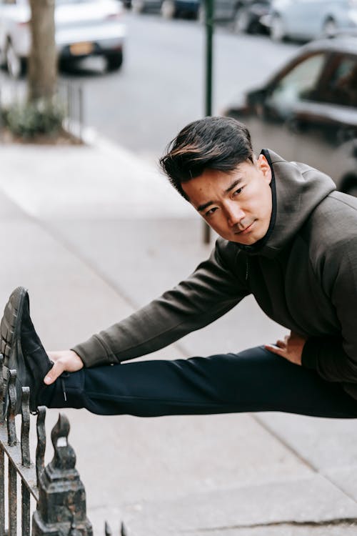 Side view of serious young ethnic male athlete with dark hair in warm activewear stretching body while leaning leg on metal fence on street during outdoor workout