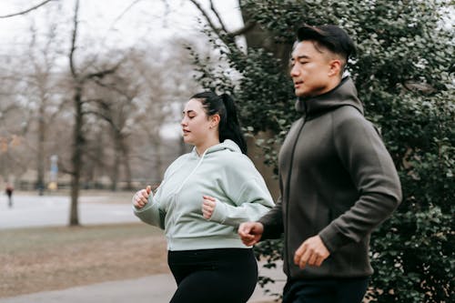 Side view of overweight woman in sportswear running with Asian trainer in city park in daytime