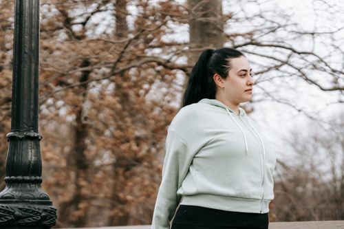 Free Side view of plus sized female with dark hair in light hoodie standing in autumn park and looking away in daytime Stock Photo