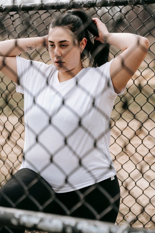Free Serious young lady in white t shirt and leggings standing near metal grid in street while working out with hands behind head in daytime Stock Photo