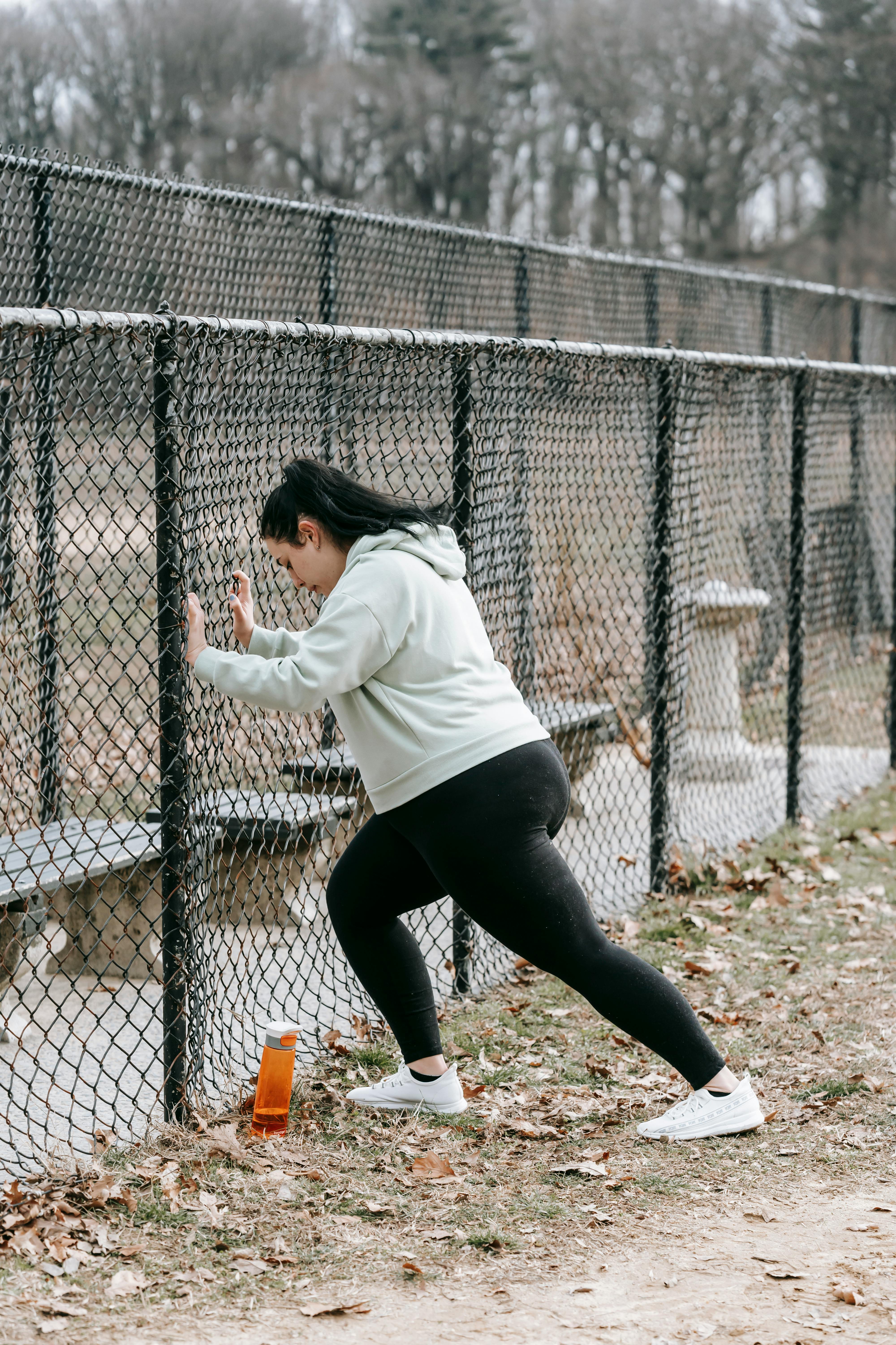 concentrated young obese woman doing exercise leaning on chain link fence in park
