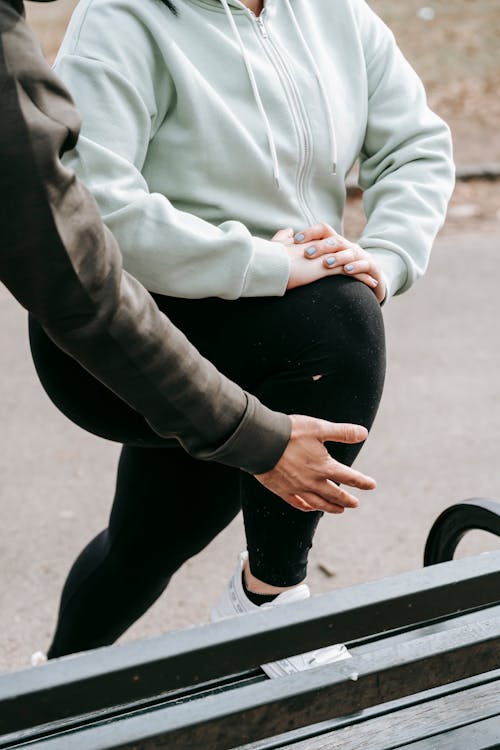 Crop unrecognizable male personal trainer helping anonymous plus size female doing stretching exercise on bench during training in park