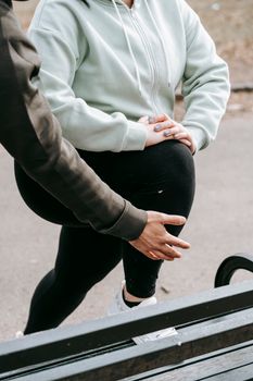 Crop unrecognizable male personal trainer helping anonymous plus size female doing stretching exercise on bench during training in park