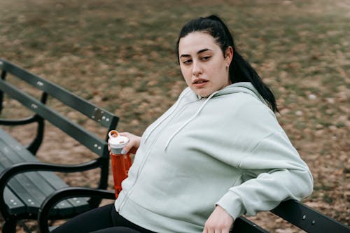 Tired young obese female with long dark hair in hoodie drinking bottle of water while resting on bench after outdoor workout in autumn park