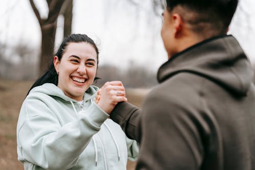 Free Cheerful plus sized female with dark hair in hoodie holding hand of unrecognizable male in park in daylight Stock Photo