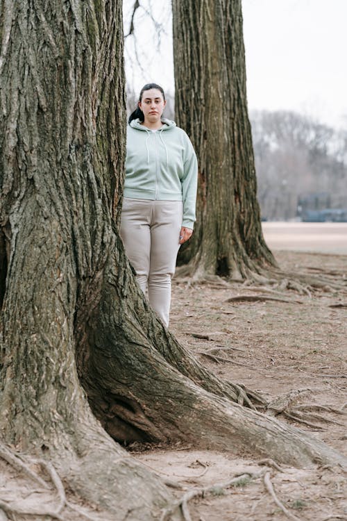 Pensive plus sized woman in activewear standing behind tree in park and looking at camera in daytime