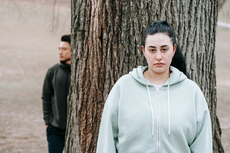 Serious Woman Before Tree And Asian Man Behind Trunk