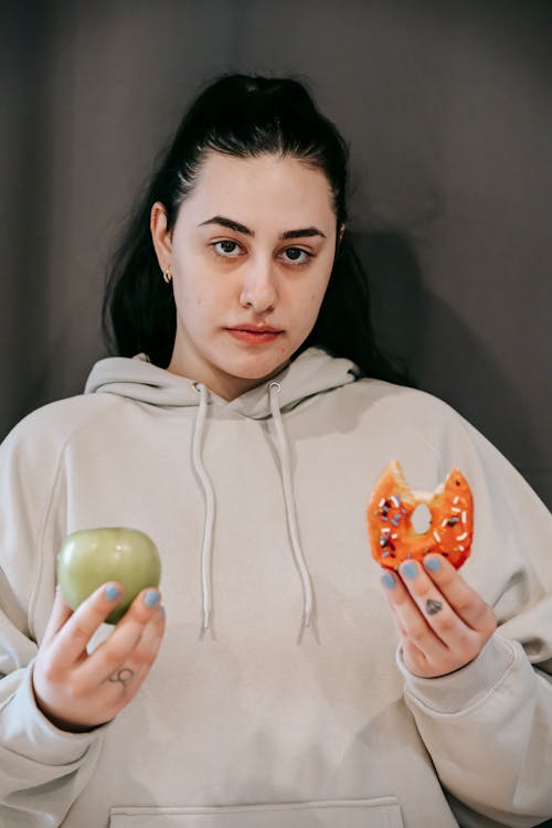 Concentrated young plus size female in sportswear looking at camera and showing healthy and junk food while making difficult choice