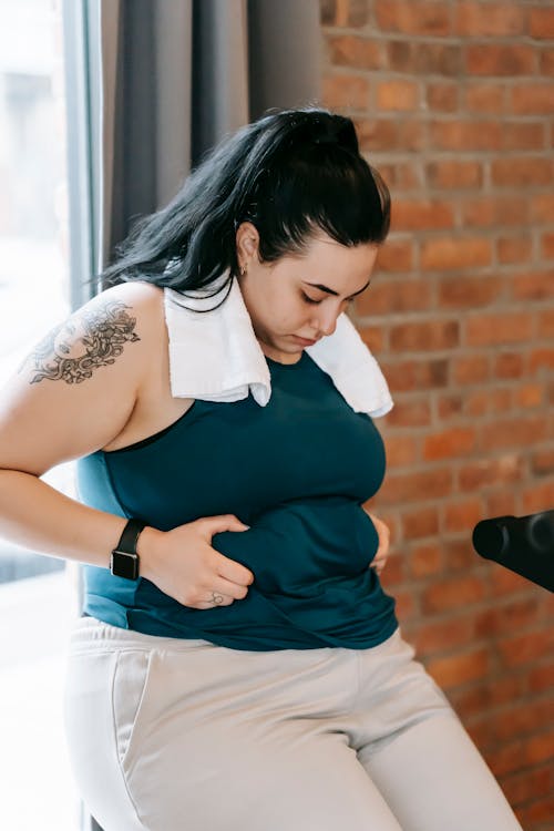 Upset young obese female in sportswear leaning on windowsill and desperately looking at stomach