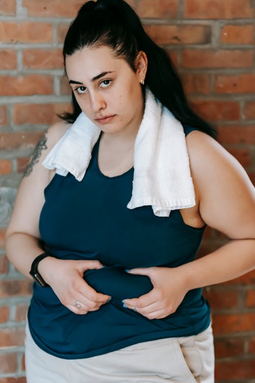 Young obese female in sportswear showing problem with overweight on belly while standing against brick wall and sadly looking at camera