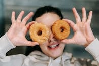 Positive young female in casual clothes smiling while covering eyes with delicious doughnuts
