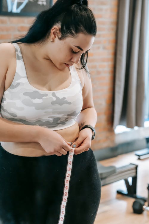 Free Young obese female using tape for evaluating size of belly while preparing for training in modern fitness center Stock Photo