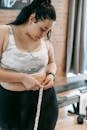 Young obese female using tape for evaluating size of belly while preparing for training in modern fitness center