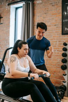 Strained overweight female in sportswear sitting on exercise machine and training with coach support
