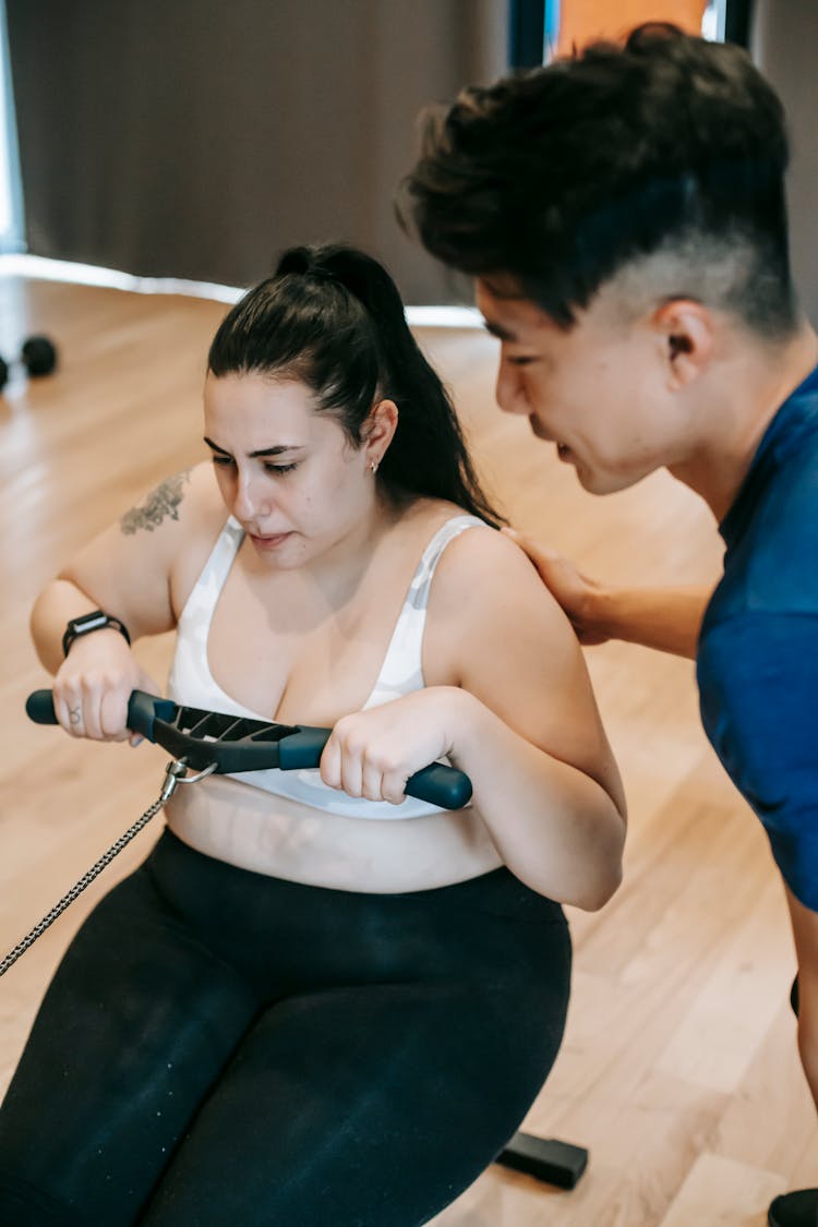 Ethnic Personal Trainer Controlling Overweight Woman Exercising On Rowing Machine