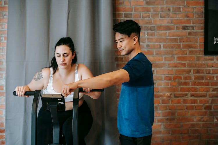 Asian Male Personal Trainer Assisting Plump Woman On Cycling Machine