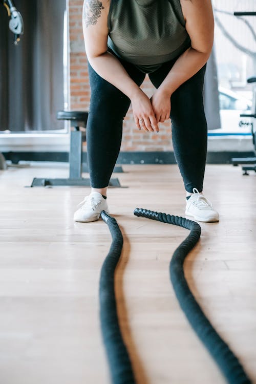 Free Crop unrecognizable plus size female in activewear leaning forward while resting after workout with battle ropes in gym Stock Photo