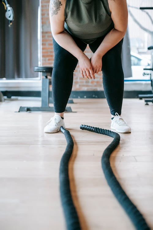 Crop unrecognizable plus size female in activewear leaning forward while resting after workout with battle ropes in gym