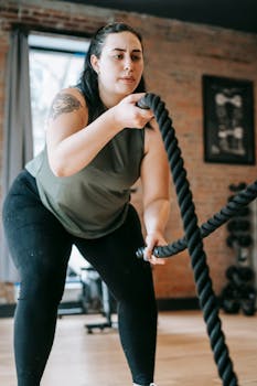 Focused plus sized female battling ropes while exercising in gym with brick walls in daytime