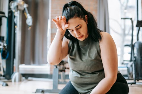 Overweight woman with dark hair in sportswear wiping sweat from face while training in gym in daytime