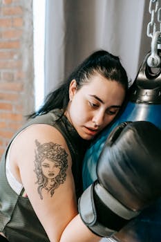 Tired female boxer with tattoo resting her head on a punching bag indoors.