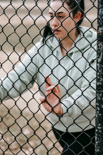 Crop concentrated female in gray hoodie leaning on net fence and looking away in daylight