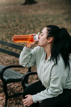 A young woman in a hoodie hydrating on a park bench during her workout break.