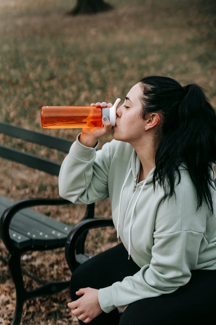 Female Jogger Drinking Water On Bench In Autumn Park