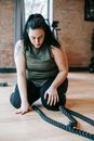 Full body concentrated plus sized female in activewear sitting on floor and exercising with battling ropes in modern gym