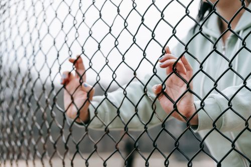 Free Crop anonymous female wearing gray hoodie touching metal net fence on street in daylight Stock Photo