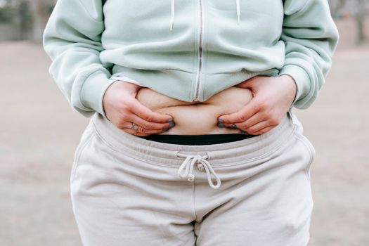 Crop anonymous plus sized female in casual hoodie touching and showing fat on belly while standing in park