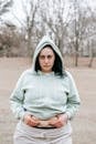 Sad plus sized female in hoodie touching and showing fat on belly while standing in leafless park and looking at camera