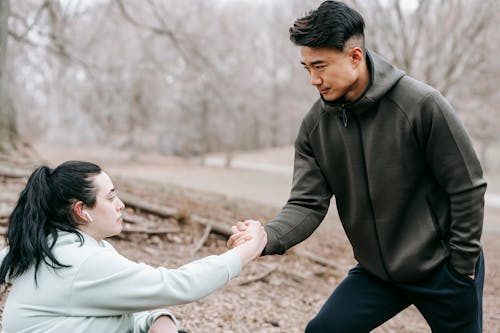 Free Young Asian male in sportswear giving hand to plump female fell on ground while training together in autumn leafless forest Stock Photo