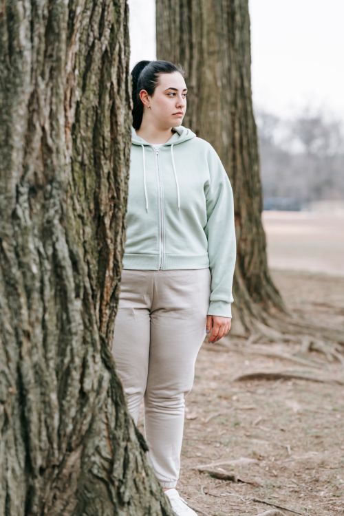 Free Sad plus sized female in light hoodie standing near tree in autumn woods on gloomy day and looking away in thoughts Stock Photo