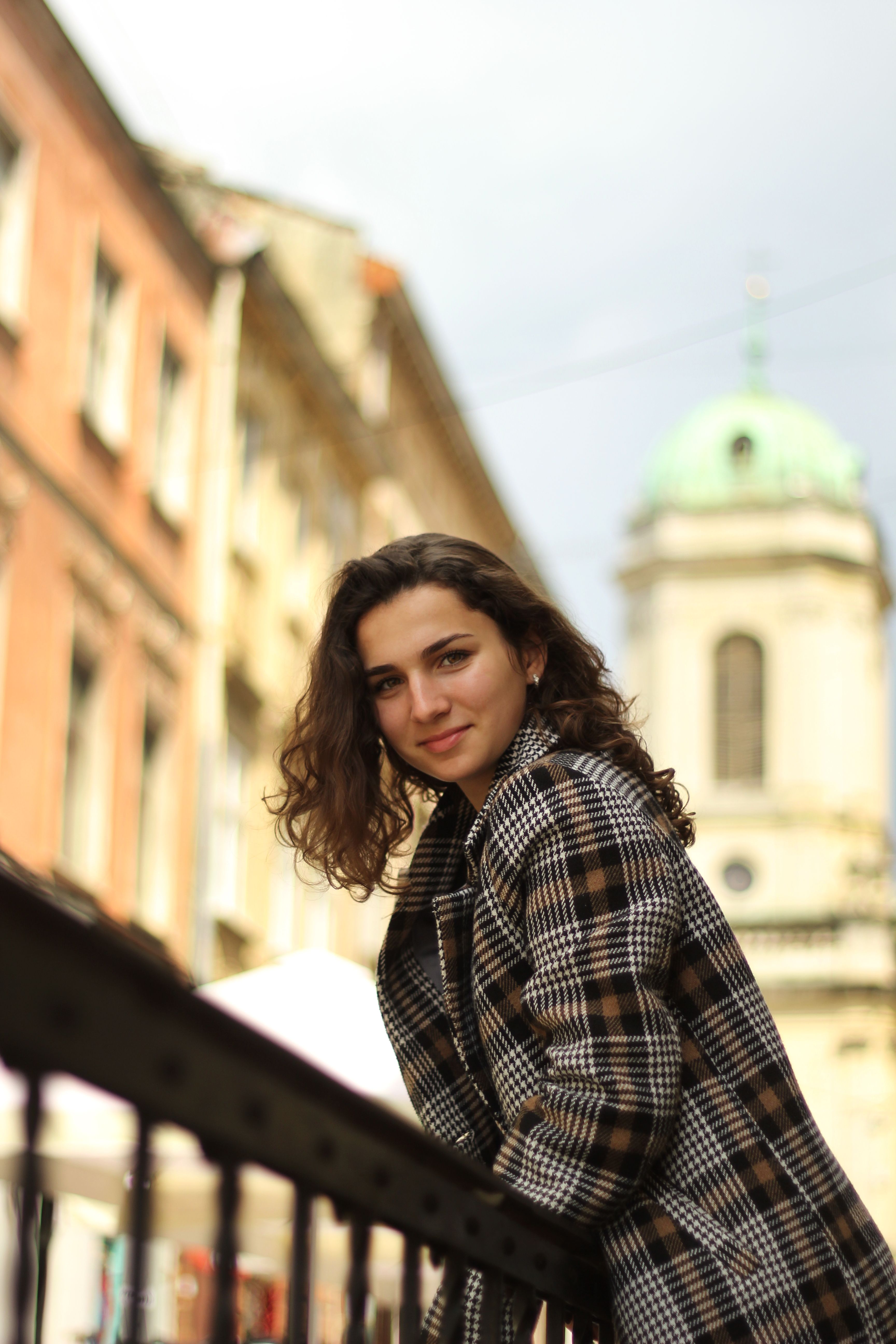 A cheerful woman in a plaid coat standing by a railing with historic architecture in the background.