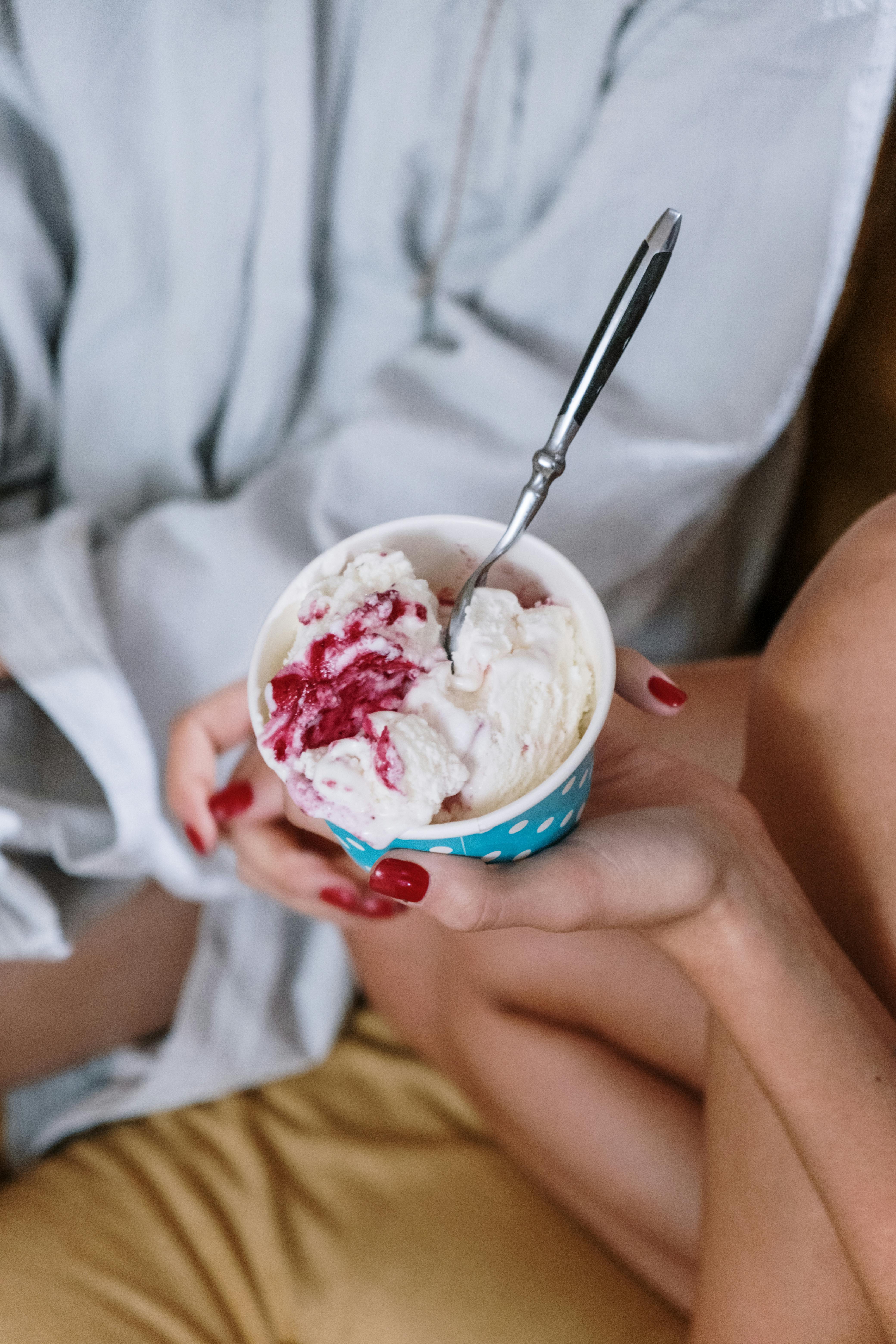 A person with manicured nails holding a cup of strawberry ice cream with a spoon, shot from above.