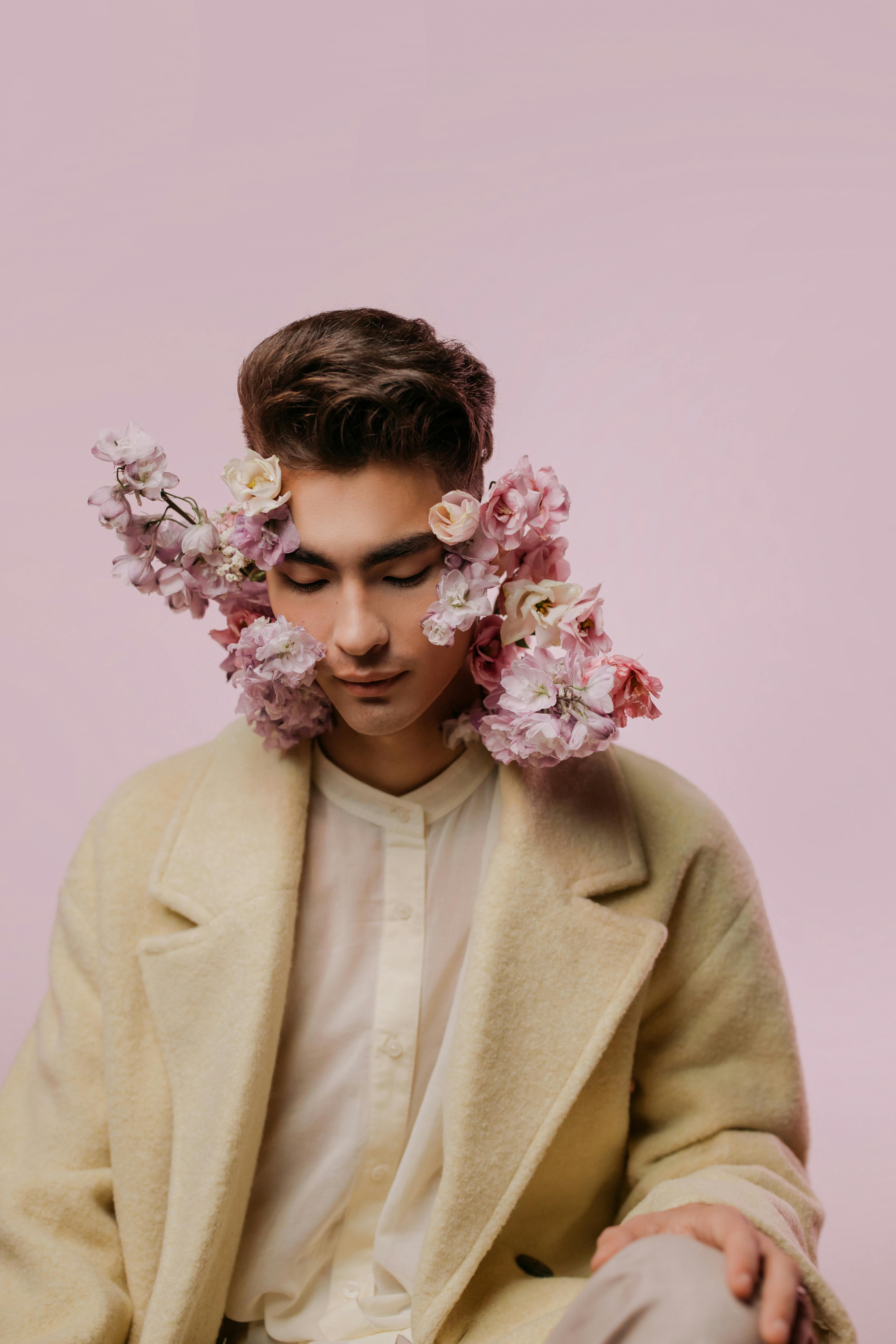 Aesthetic portrait of a man adorned with pastel pink flowers, wearing a beige coat, in a studio setting.