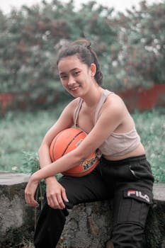 Portrait of a young woman in casual attire holding a basketball outdoors.