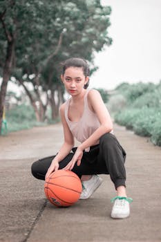 Portrait of a young woman kneeling with a basketball on an outdoor pathway.
