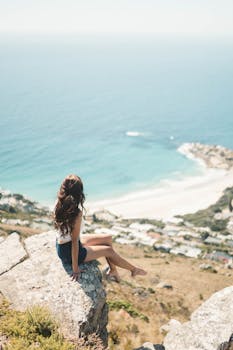 A serene scene of a woman sitting on a cliff with a panoramic view of Cape Town's stunning coastline.
