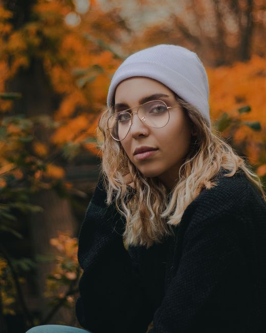 Side view of young emotionless lady in casual clothes and hat with glasses sitting near orange foliage on plants and trees in nature and looking at camera in fall