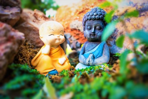 Free Miniature Buddha and monk statues sit peacefully amidst lush garden flora. Stock Photo