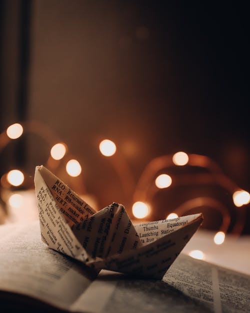 Paper ship placed on book near glowing garland in room